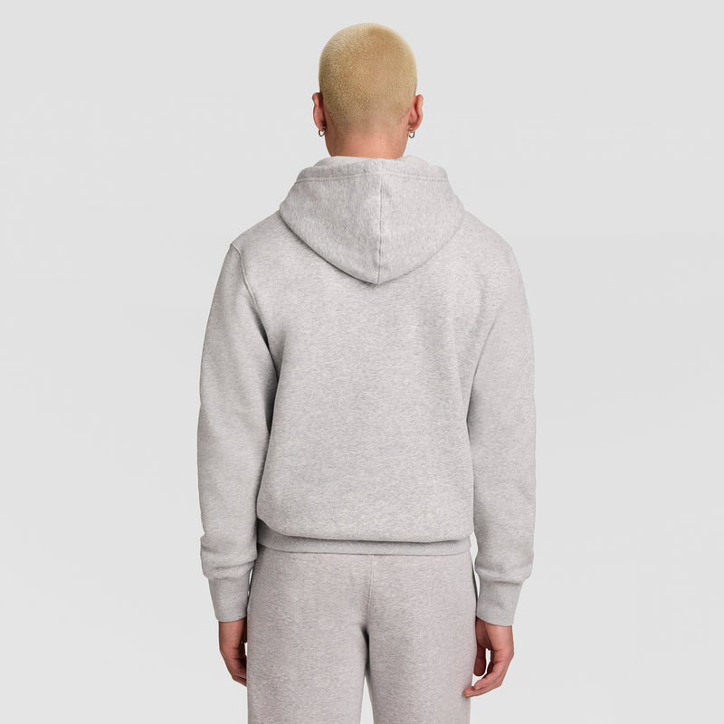 A person with short blond hair, wearing a light gray Champion Powerblend Hoodie featuring the full script logo and matching sweatpants, is standing with their back to the camera against a plain light background.