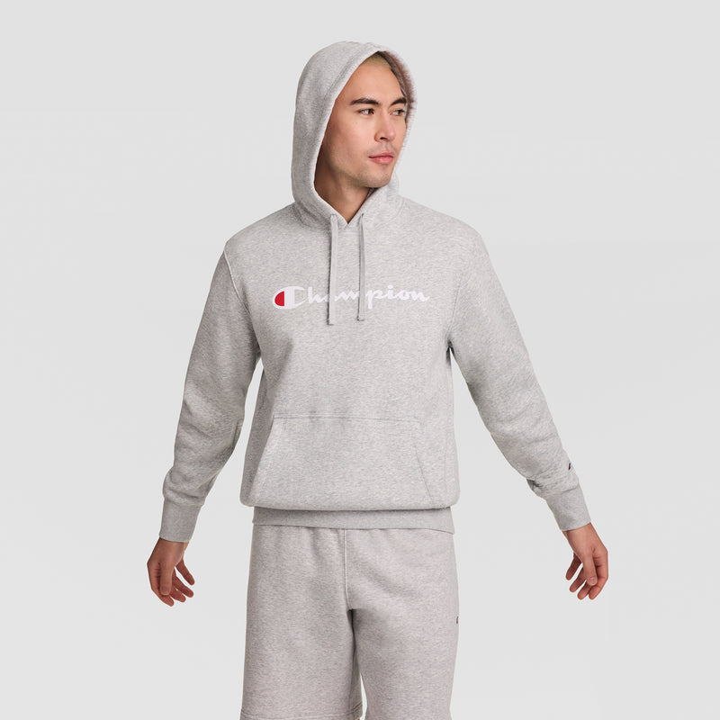 A person wearing a light gray Champion Powerblend Hoodie with the Full Script Logo and matching sweatpants stands with arms slightly out, looking to the side against a plain light background.