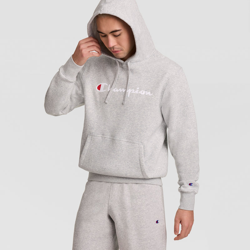 A man wears a light gray Champion Powerblend Hoodie with the Full Script Logo and matching sweatpants, hood up, looking down and adjusting the hood with one hand against a plain background.