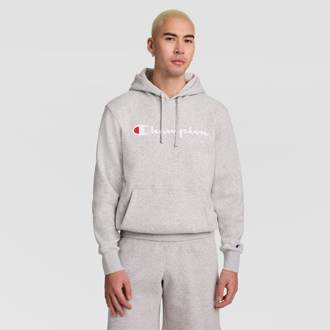 A person with short blond hair wears a light gray Champion Powerblend Hoodie with a full script logo and matching sweatpants, standing against a plain light gray background.