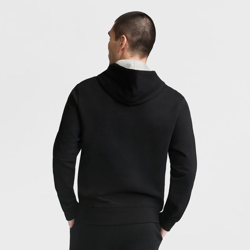 A man with short hair is shown from the back, wearing a black Champion Powerblend Hoodie with a Full Script Logo and black pants, standing against a plain white background.