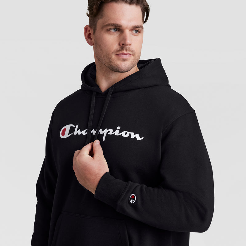 A man in a black Champion Powerblend Hoodie, Full Script Logo - Big & Tall, stands against a light background, looking to the side with one hand holding the collar. The Champion logo is displayed across his chest and on his sleeve.