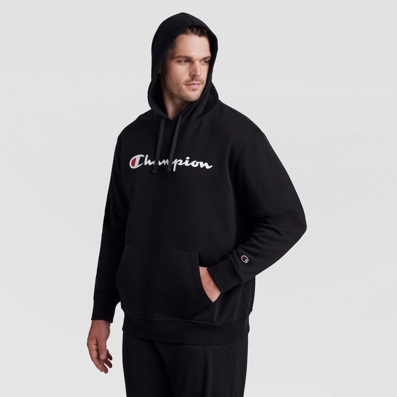 A man wears the Champion Powerblend Hoodie, Full Script Logo - Big & Tall, featuring a white and red logo on the chest, black fleece fabric, and hood up. He stands against a light gray background with one hand in his pocket.