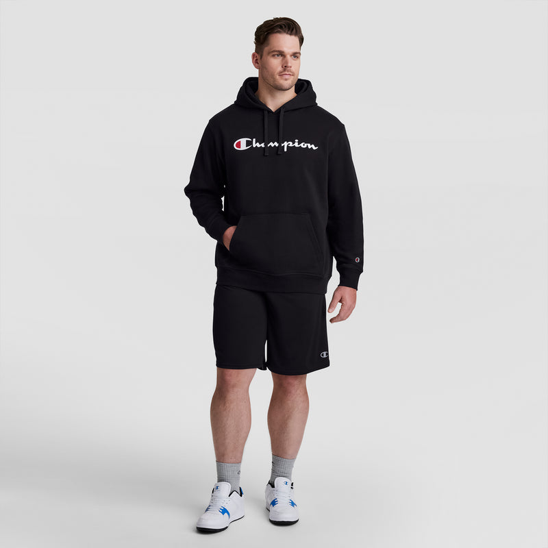 A man wears a black Champion Powerblend Hoodie, Full Script Logo - Big & Tall, with matching black shorts, gray socks, and white sneakers, posed against a plain light gray background.