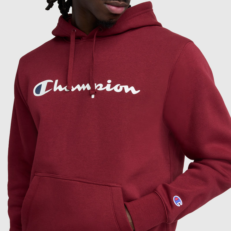 A person wearing a maroon Champion Powerblend Hoodie, Full Script Logo, featuring the brand's logo across the chest and a small C logo on the sleeve, stands against a plain light gray background.