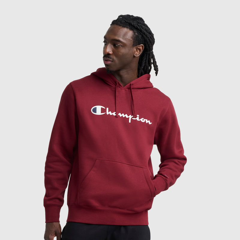 A man with long braided hair wears a maroon Champion Powerblend Hoodie, Full Script Logo, and black pants while standing against a plain light gray background, looking left with one hand in his pocket.