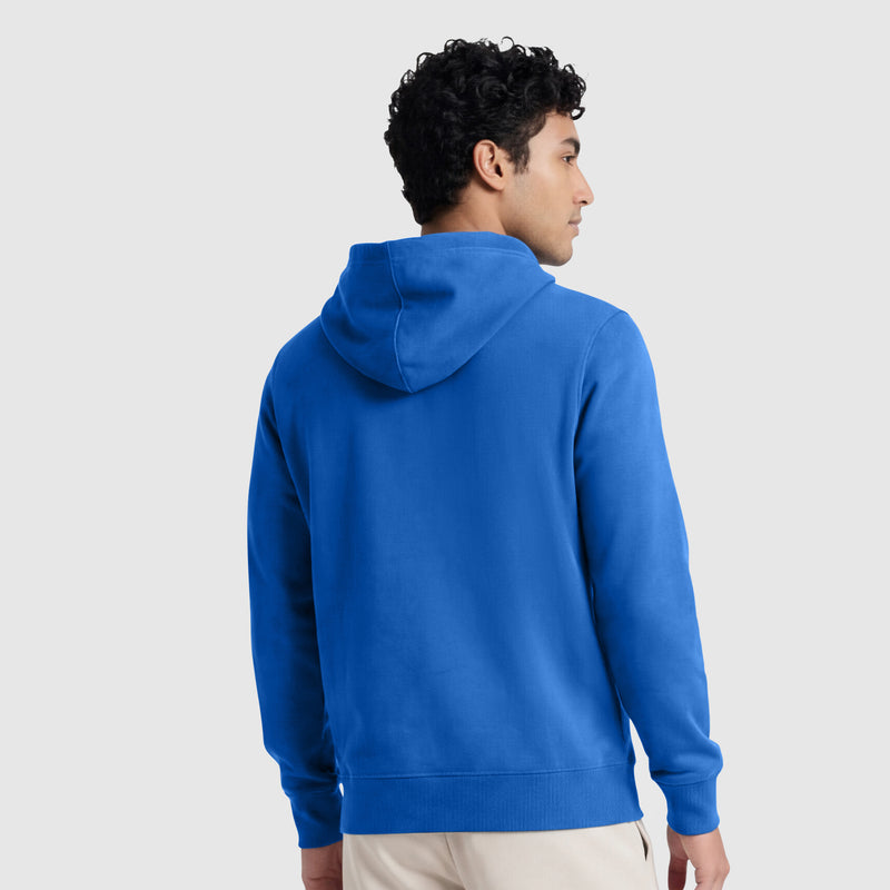 A person with short curly hair is wearing a bright blue Champion Powerblend Hoodie, Full Script Logo, and beige pants, facing away from the camera against a plain light grey background.