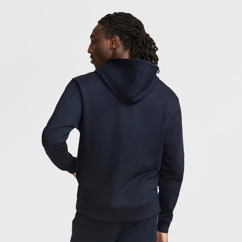 A person with medium-length curly hair is seen from the back, wearing a dark navy Champion Powerblend Hoodie with Full Script Logo and matching pants, standing against a plain white background.