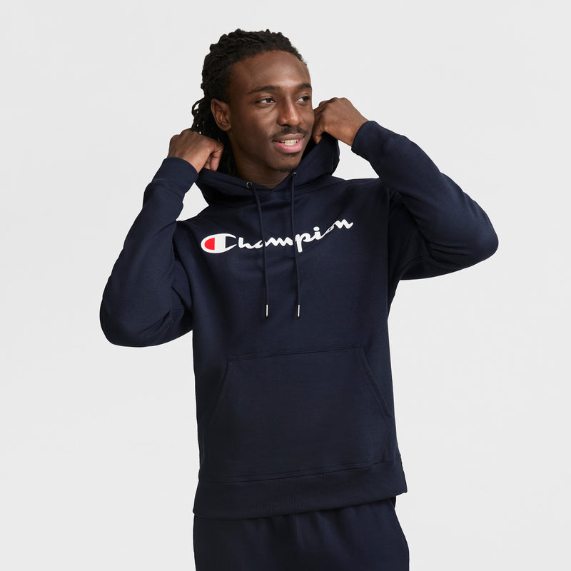 A man smiles while adjusting the hood of a navy blue Champion Powerblend Hoodie, Full Script Logo, standing against a plain white background.
