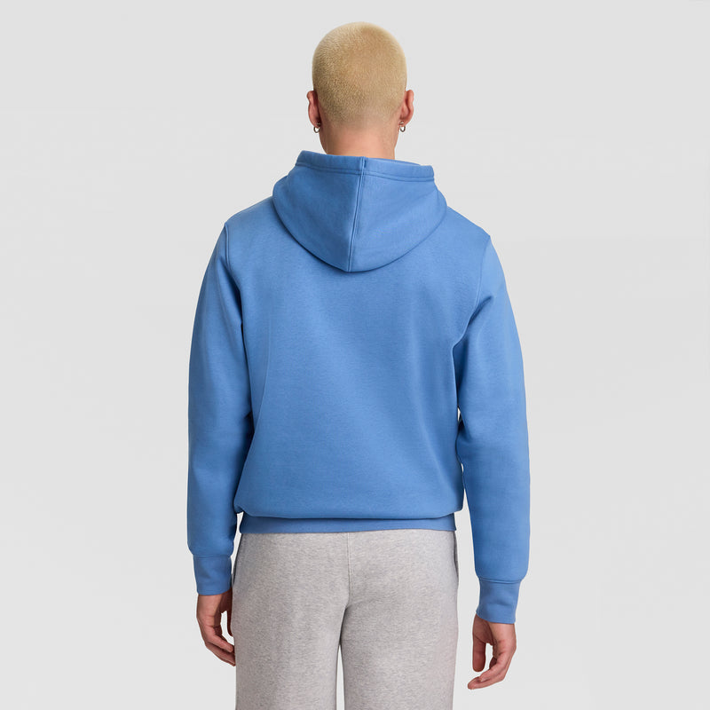 A person with short blond hair stands with their back to the camera against a plain light background, wearing a Champion Powerblend Hoodie, Full Script Logo in blue and light gray pants.