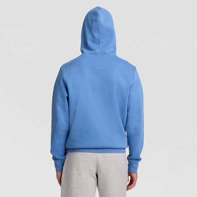 A person wearing the Champion Powerblend Hoodie, Full Script Logo in blue and light gray sweatpants stands facing away from the camera against a plain light background.