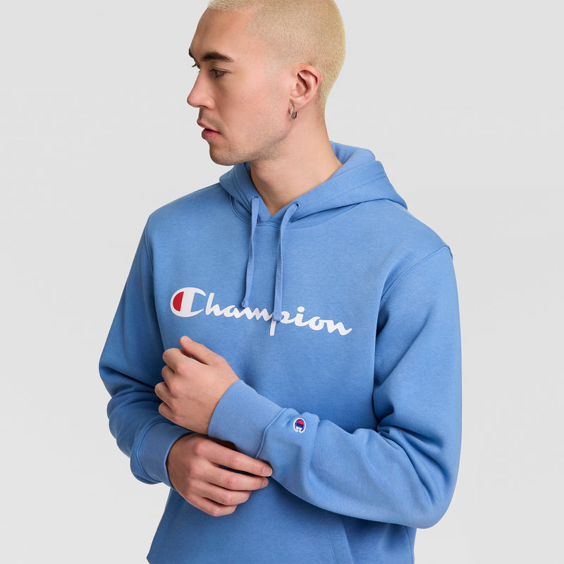 A person with short blond hair, wearing the Champion Powerblend Hoodie with a Full Script Logo in blue, stands against a plain light background, looking to the side with one arm crossed over their chest.
