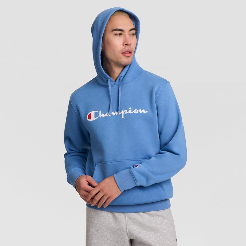 A person wearing a blue Champion Powerblend Hoodie, Full Script Logo, with the hood up and gray sweatpants looks to the side against a plain light background.