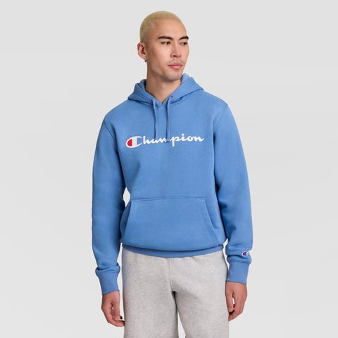 A person with short blond hair wears a blue Champion Powerblend Hoodie, Full Script Logo, paired with light gray sweatpants. They stand against a plain light background, looking slightly to the side with their hands relaxed by their sides.