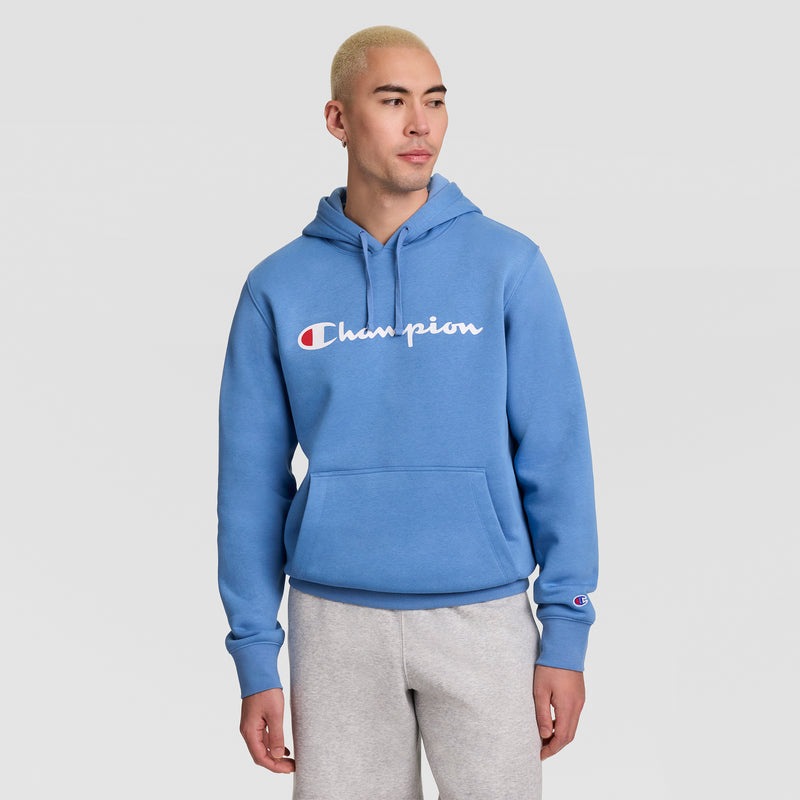 A person with short blond hair wears a blue Champion Powerblend Hoodie, Full Script Logo, paired with light gray sweatpants. They stand against a plain light background, looking slightly to the side with their hands relaxed by their sides.