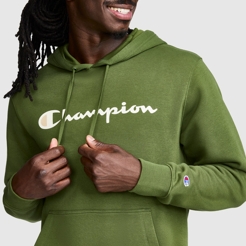 A person wearing a green Champion Powerblend Hoodie with the Full Script Logo smiles while adjusting the drawstrings. Only part of their face and upper body are visible, highlighting the cozy style of this classic Champion hoodie.