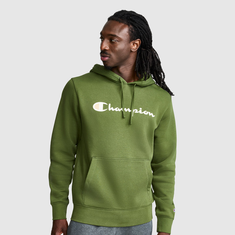 A man wearing a green Champion Powerblend Hoodie, Full Script Logo, with a front pocket and gray pants stands against a plain light gray background, looking to his left.