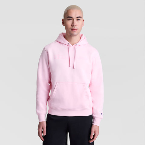 A person with short blond hair wears a light pink Champion Powerblend Hoodie with a tonal script logo and black pants, standing facing forward against a plain light gray background.
