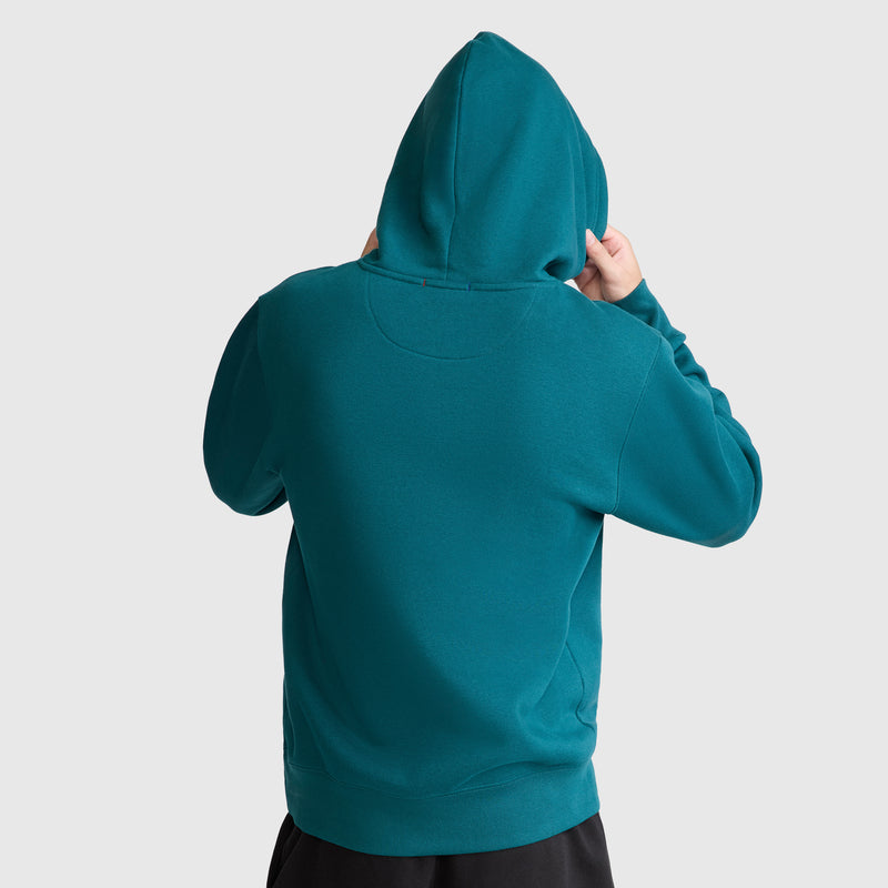 A person wearing a teal Champion Powerblend Hoodie with a tonal script logo stands facing away from the camera, both hands adjusting the hood. The plain, light background emphasizes the comfort of the Powerblend Fleece fabric.