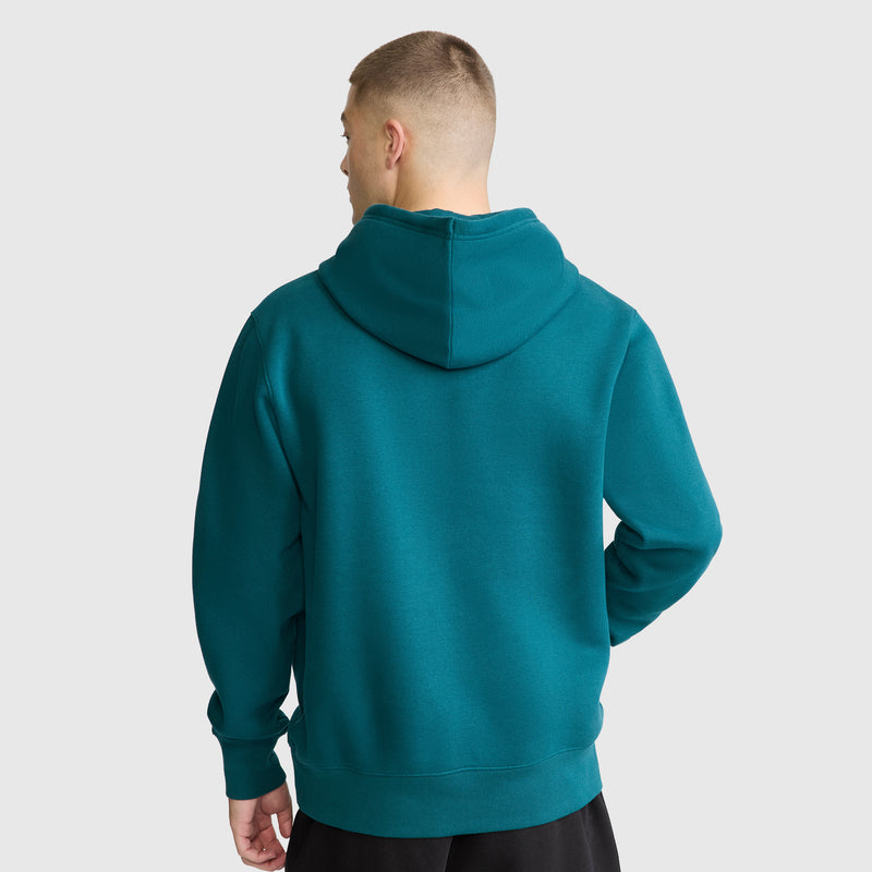 A person with short hair, wearing a teal Champion Powerblend Hoodie with a tonal script logo and black pants, stands facing away against a plain light gray background.