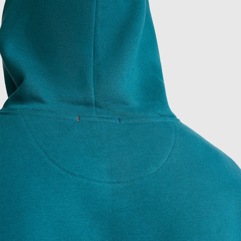 Close-up of the back of a teal Champion Powerblend Hoodie, Tonal Script Logo, featuring the hood and upper back. Two small thread stitches—one red and one blue—stand out near the seam against a plain light gray background. This design subtly emphasizes comfort while honoring Champion’s classic script logo heritage.