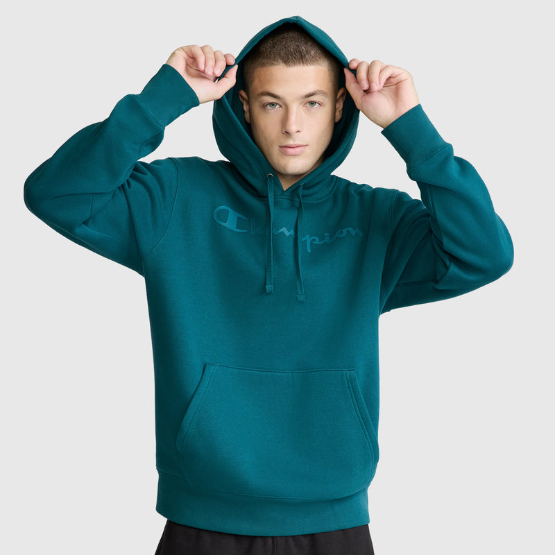 A person wearing the Champion Powerblend Hoodie with a tonal script logo on the front holds the hood with both hands and looks at the camera, standing against a plain light background.