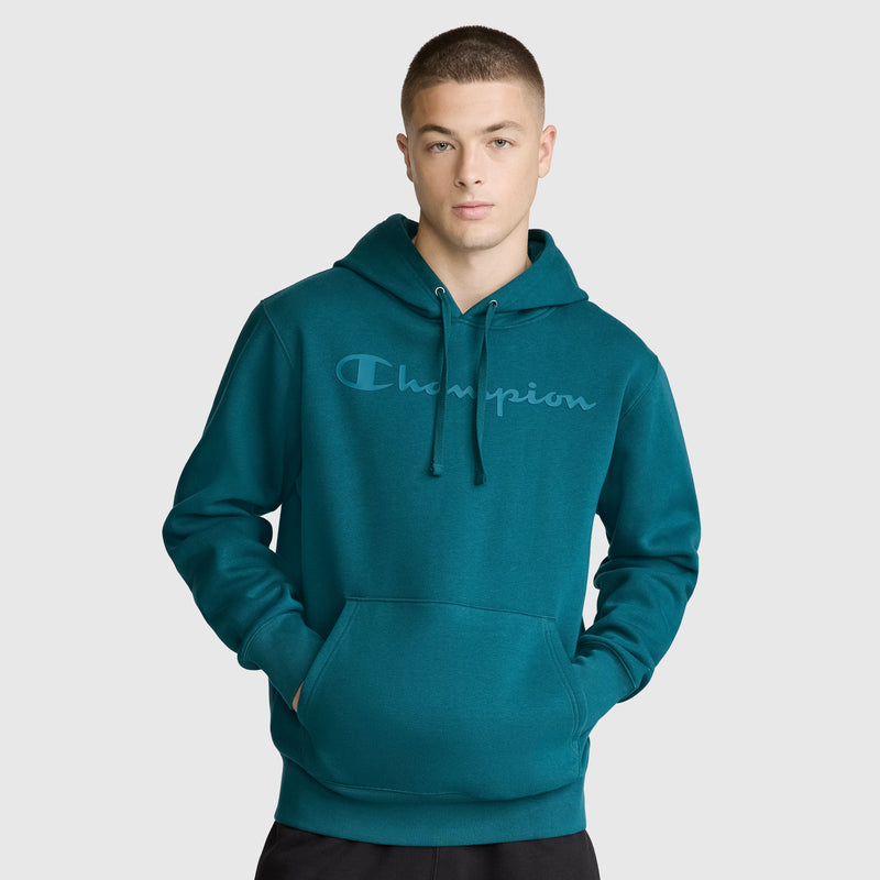 A person wearing a teal Champion Powerblend Hoodie with a tonal script logo and front pocket stands against a plain light gray background, looking at the camera with their hands in the pocket.