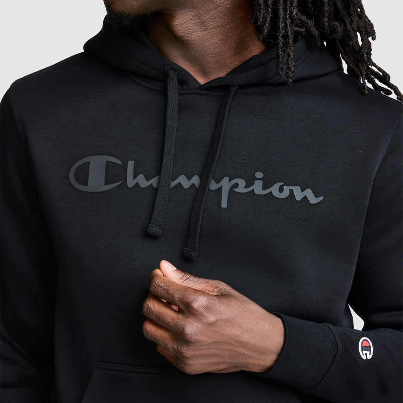 A person wearing a black Champion Powerblend Hoodie with a tonal script logo across the chest and a small Champion logo on the sleeve, holding part of the hoodie with one hand.