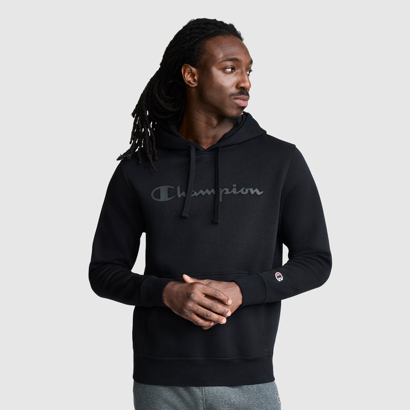 A man with long dreadlocks wears a black Champion Powerblend Hoodie, Tonal Script Logo, paired with gray sweatpants. He stands against a plain light gray background, looking to his right with his hands clasped.