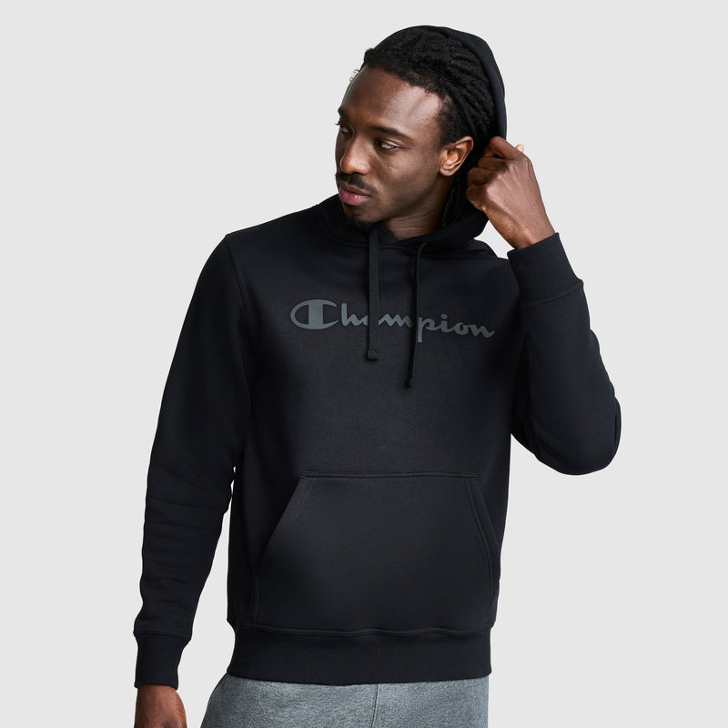 A man wearing a black Champion Powerblend Hoodie with a tonal script logo and gray pants adjusts his hood while looking to the side against a plain light background.