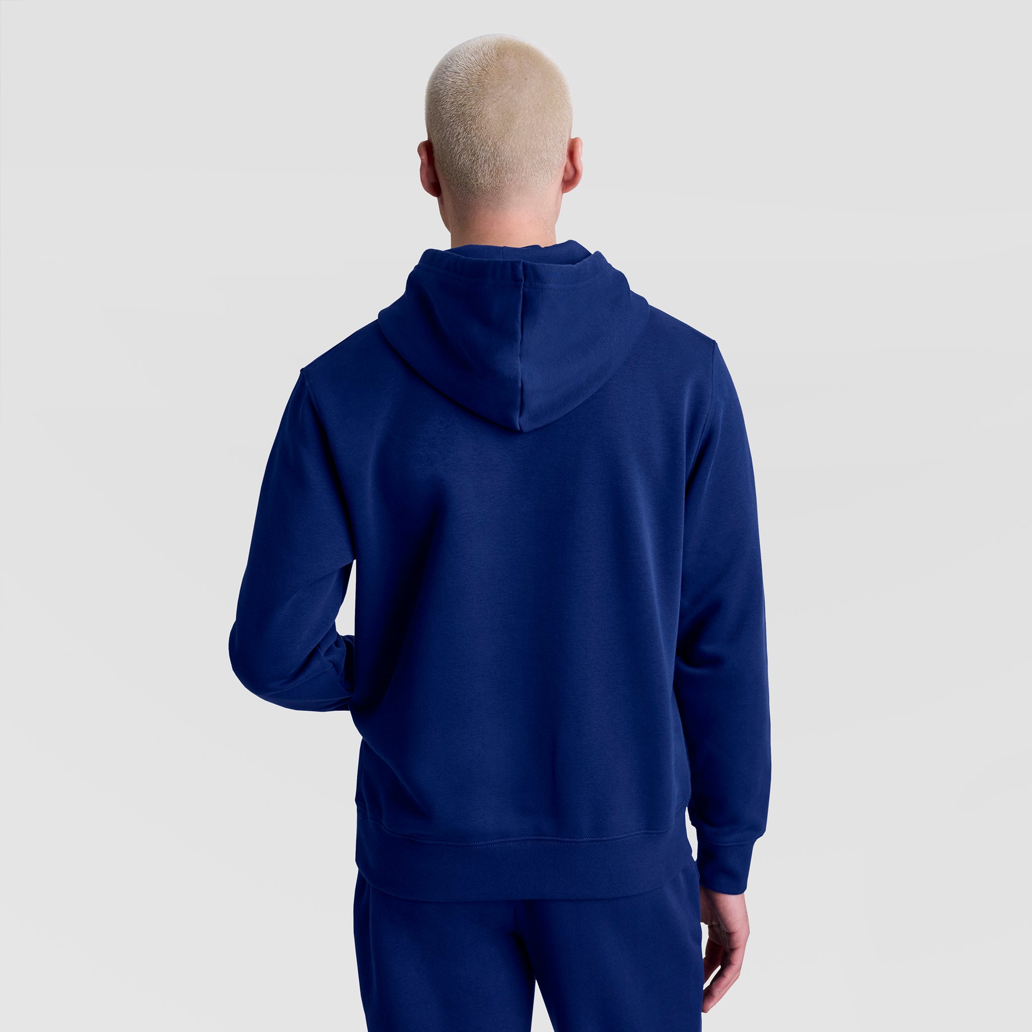 A person with short, bleached blonde hair is shown from the back in a royal blue Champion Powerblend Hoodie, Tonal Script Logo, and matching pants against a plain light gray background.
