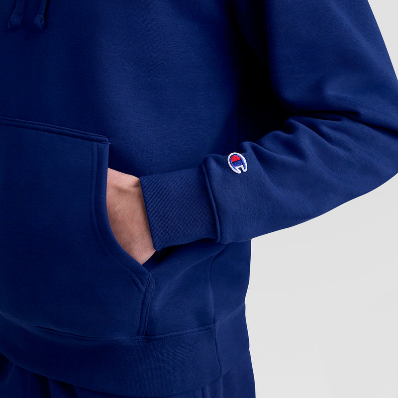 A person wears a blue Champion Powerblend Hoodie, Tonal Script Logo, with one hand in the front pocket. The tonal script logo patch is visible on the sleeve. The background is plain and light-colored.