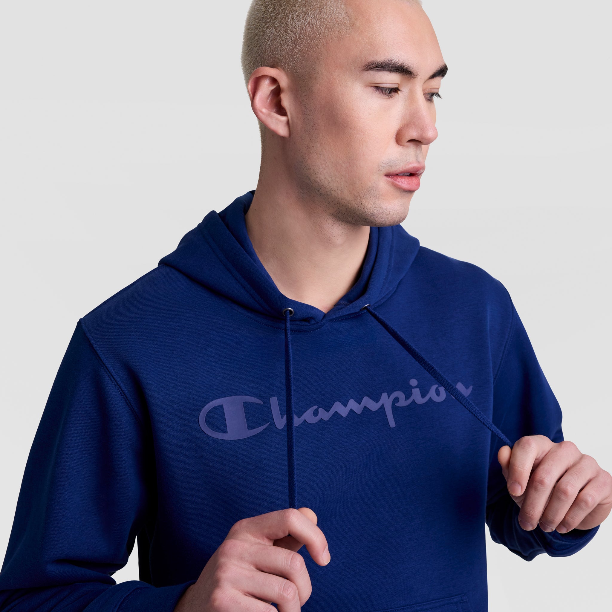 A person in a blue Champion Powerblend Hoodie with a tonal script logo looks down and holds the drawstrings. The plain, light background highlights the cozy design of this men's fleece hoodie.