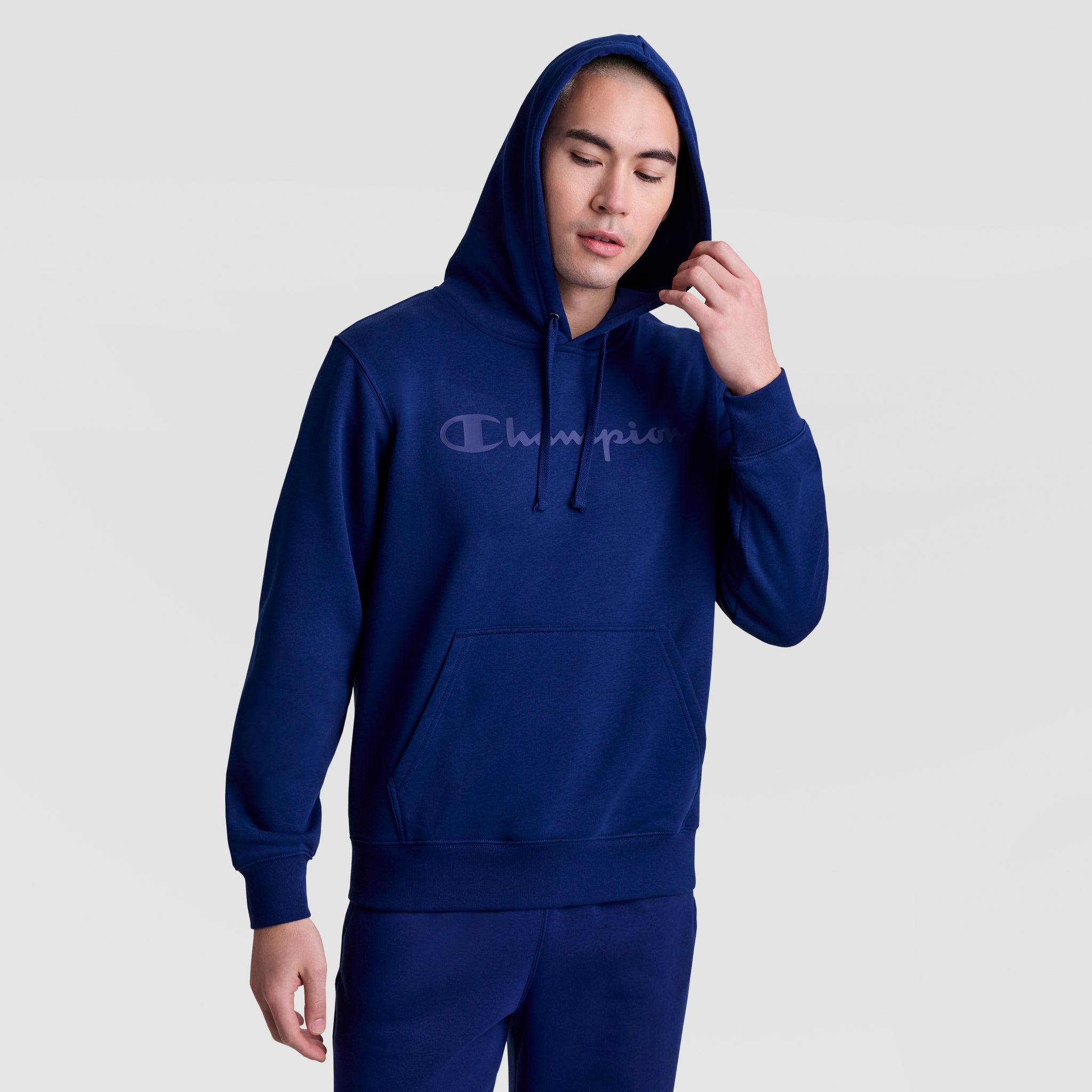 A man in a Champion Powerblend Hoodie, Tonal Script Logo, stands against a light gray background, holding the men's hoodie drawstring near his head with eyes closed, wearing matching pants.