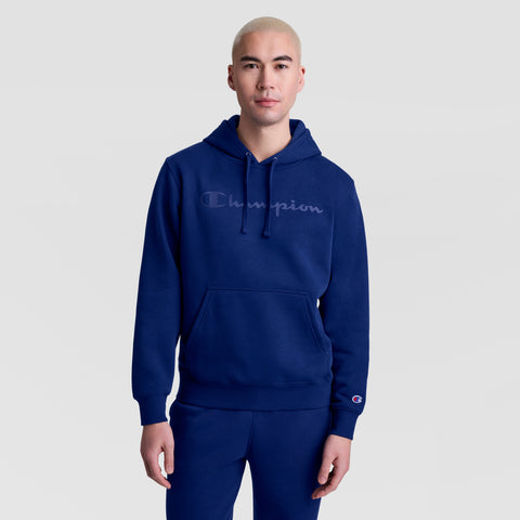 A person with short, light hair wears a blue Champion Powerblend Hoodie with a tonal script logo and matching blue pants, standing against a plain light gray background.