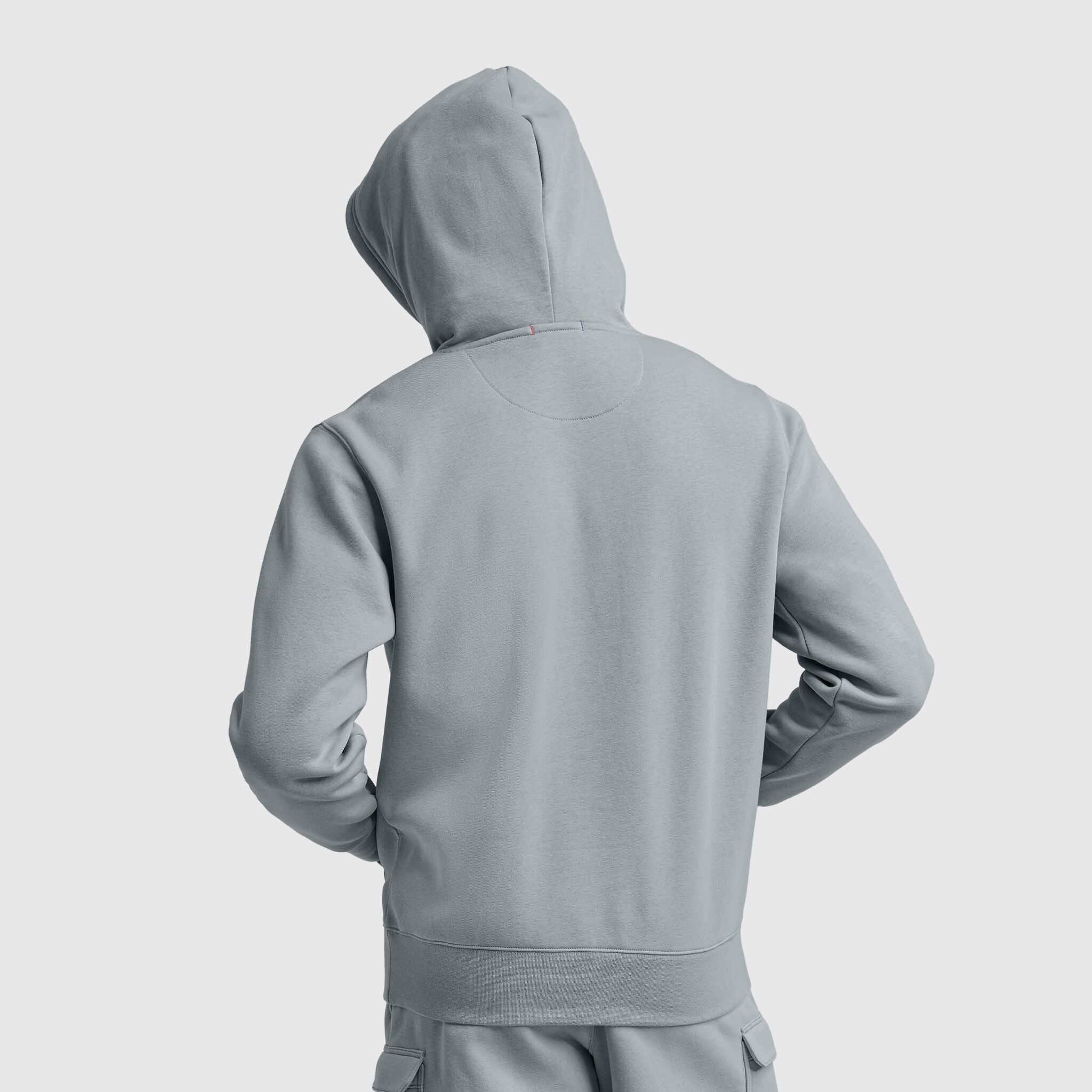 A person stands with their back to the camera, hood up and hands on hips, wearing the Champion Powerblend Hoodie with a tonal script logo in light gray against a plain background.