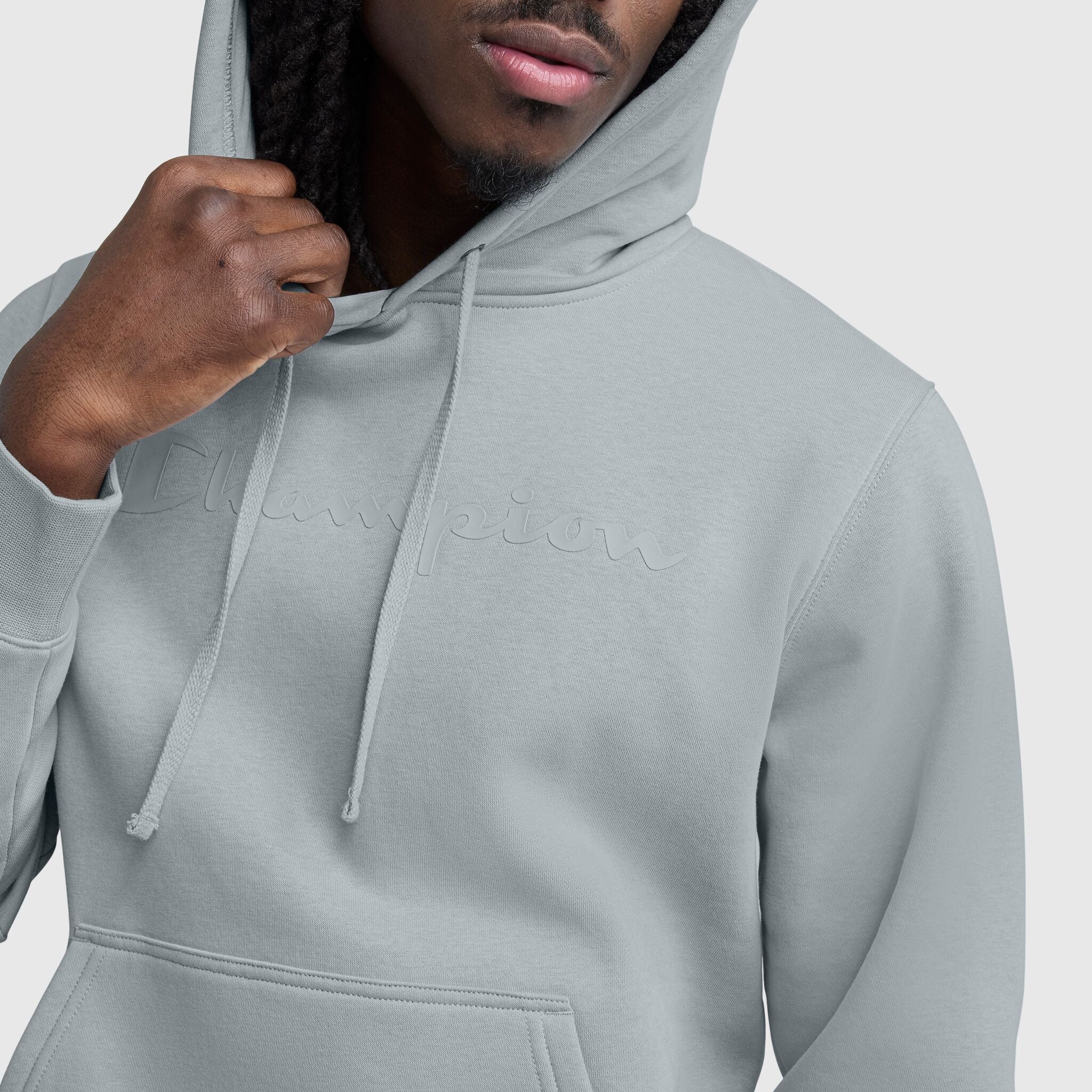 A person wears a light gray Champion Powerblend Hoodie, Tonal Script Logo, with the hood up and holding a drawstring, showing only the lower half of their face. The tonal logo adds a sleek touch to this men's hoodie by Champion.