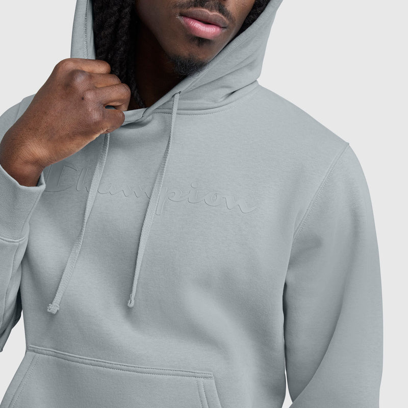 A person wears a light gray Champion Powerblend Hoodie, Tonal Script Logo, with the hood up and holding a drawstring, showing only the lower half of their face. The tonal logo adds a sleek touch to this men's hoodie by Champion.