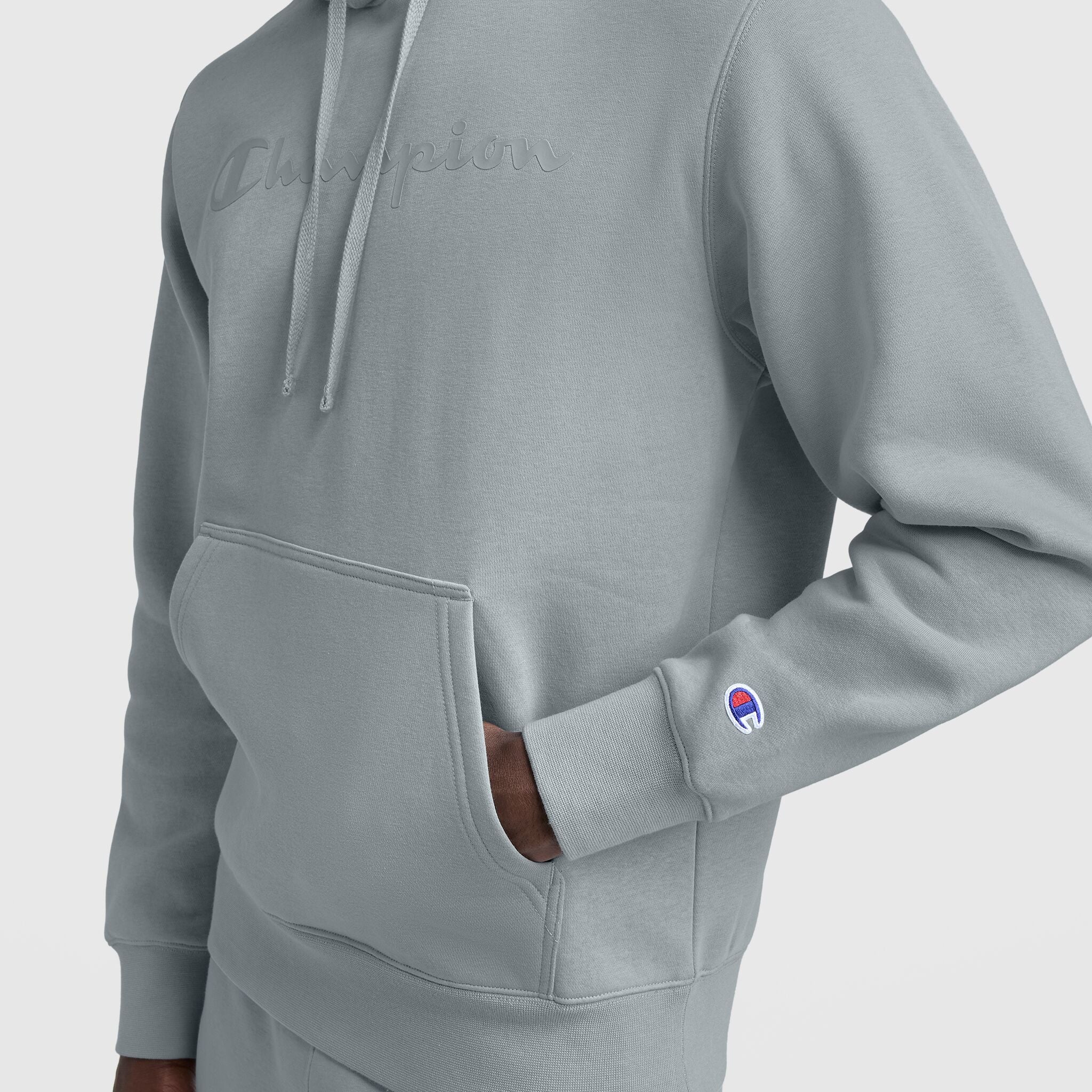 A person wears a light gray Champion Powerblend Hoodie with Tonal Script Logo, one hand in the front pocket. The embossed chest logo and small sleeve emblem stand out against a plain white background.