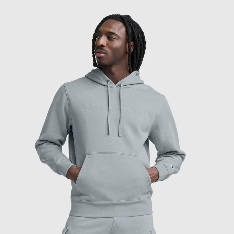 A man with shoulder-length dreadlocks wears a light gray Champion Powerblend Hoodie with a tonal script logo and matching sweatpants, standing hands-in-pocket against a plain light gray background.