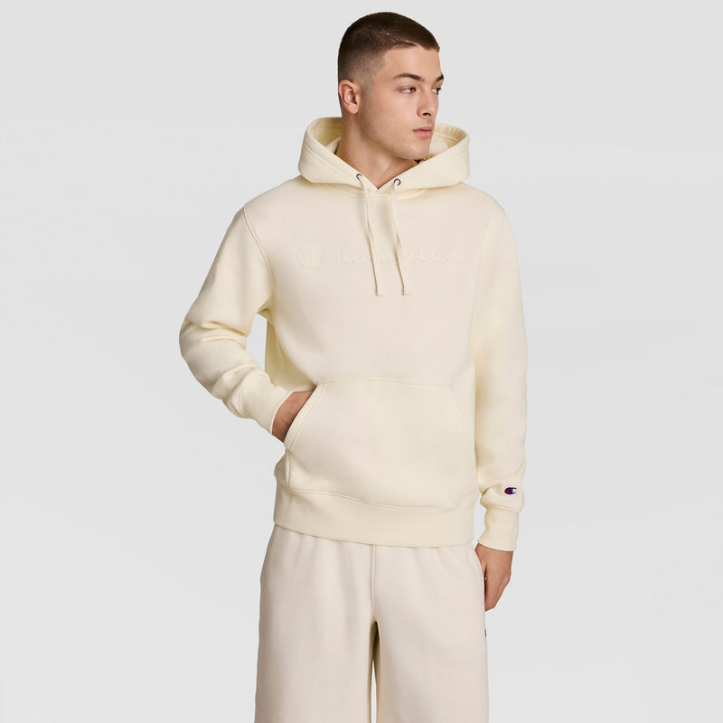 A young man wears a cream-colored Champion Powerblend Hoodie with a tonal script logo and matching sweatpants, standing with one hand in his pocket and looking to the side. The hoodie includes a front pocket and features classic detailing.