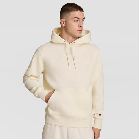 A man wears the Champion Powerblend Hoodie, Tonal Script Logo, an off-white sweatshirt with a front pocket and subtle embroidery. He stands with one hand in the pocket, looking to the side. Soft Powerblend Fleece ensures comfort.