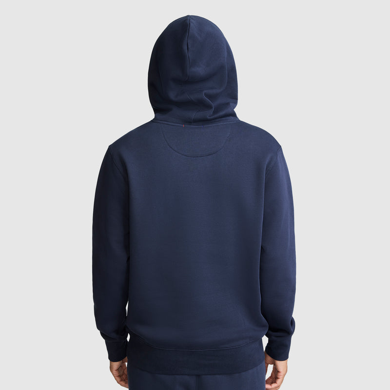 A person is seen from the back wearing a navy blue Champion Powerblend Hoodie with a tonal script logo, standing against a plain light gray background. The hood is up and their hands are relaxed by their sides, highlighting the comfortable fit of this Powerblend Fleece.