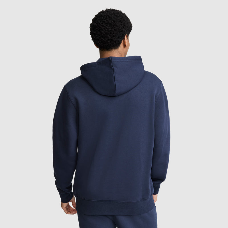 A person stands facing away, wearing a Champion Powerblend Hoodie with a tonal script logo in navy blue and matching navy pants. The hood is down, and the plain light gray background highlights the hoodie’s classic style.