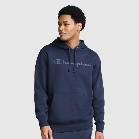 A man wearing a navy blue Champion Powerblend Hoodie with a tonal script logo, styled with matching sweatpants, stands against a plain light gray background and looks off to the side.