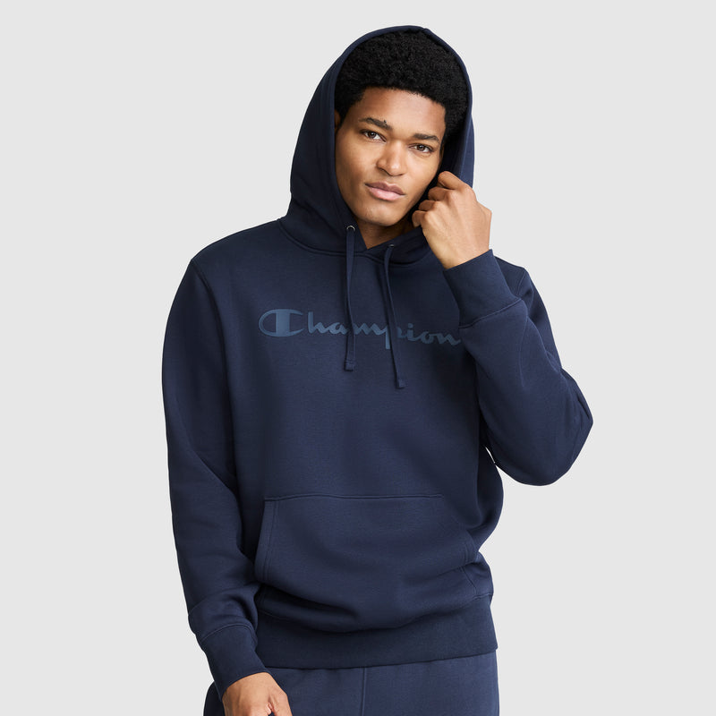 A person wearing a navy blue Champion Powerblend Hoodie with a tonal script logo and front pocket stands against a plain light gray background, touching the side of their hood and looking at the camera.