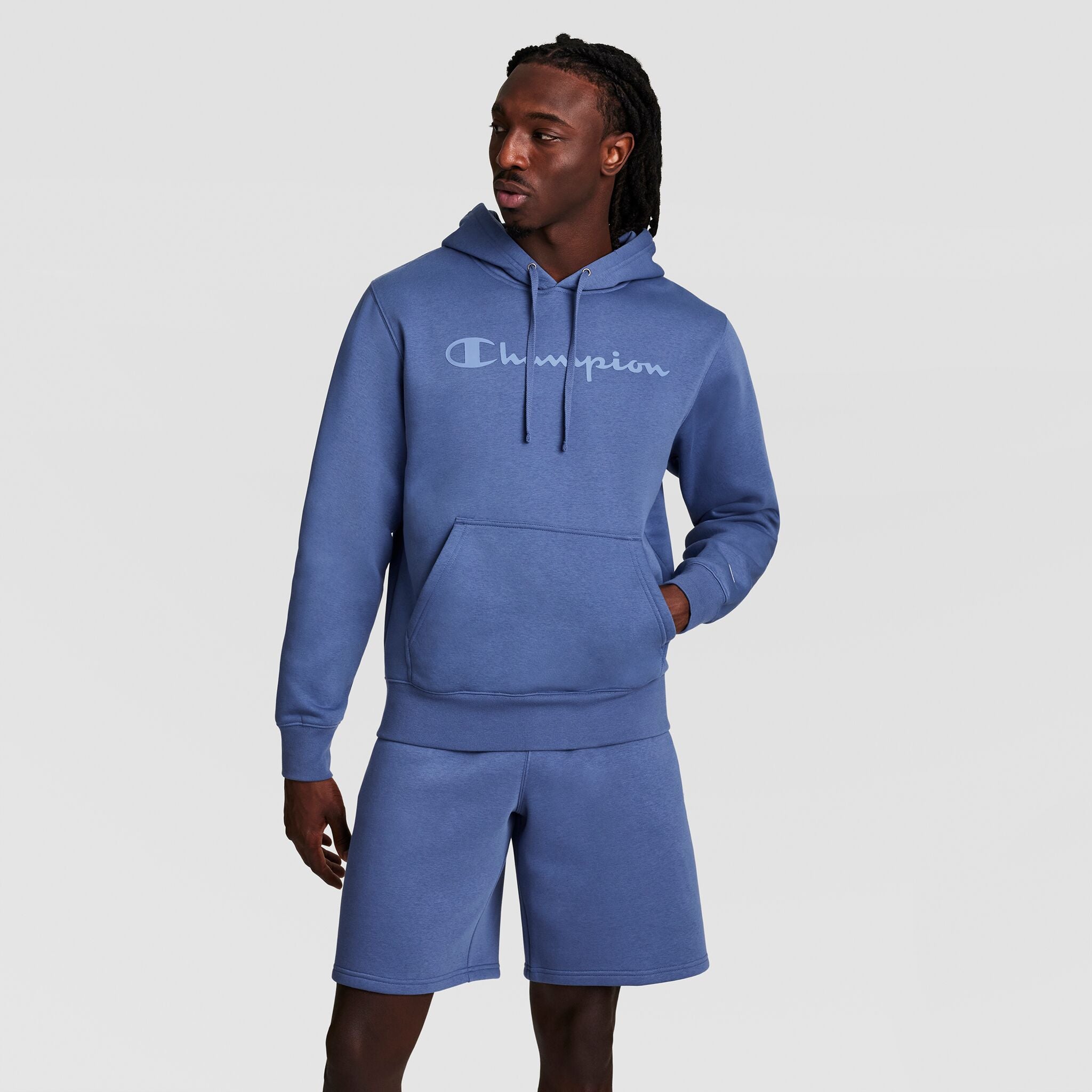 A man in a blue Champion Powerblend Hoodie with a tonal script logo and matching shorts stands with one hand in his pocket against a plain light gray background.