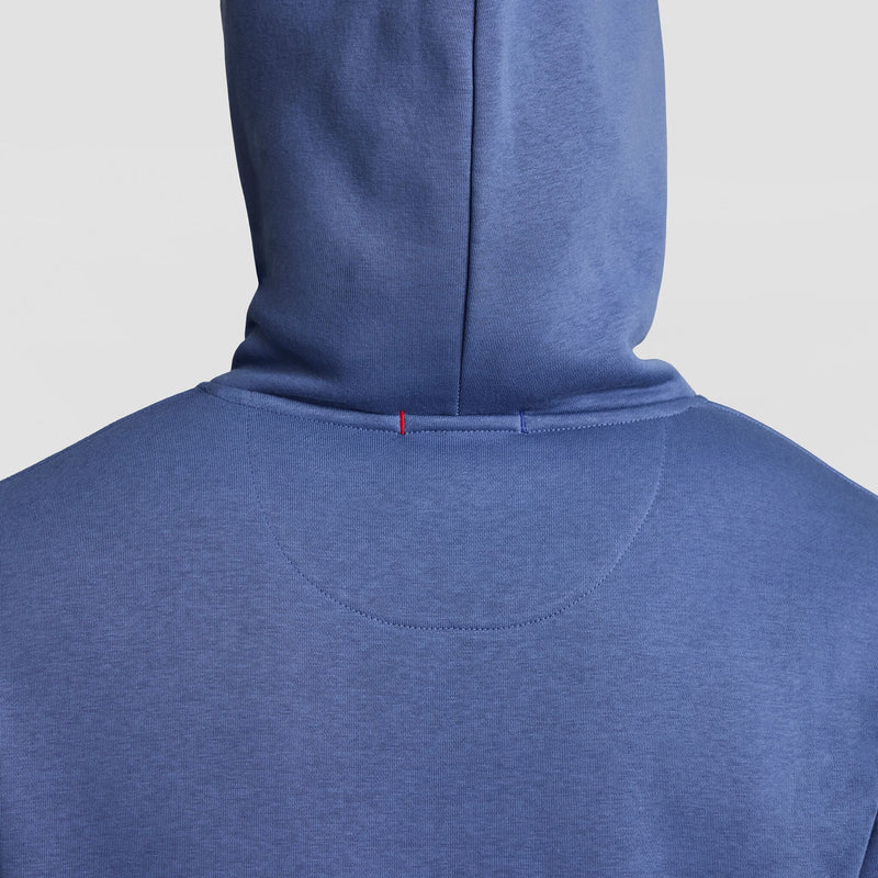 The image shows the back of a person wearing a blue Champion Powerblend Hoodie, Tonal Script Logo, with the hood up. A small red stitched detail appears near the neckline. The background is plain white.