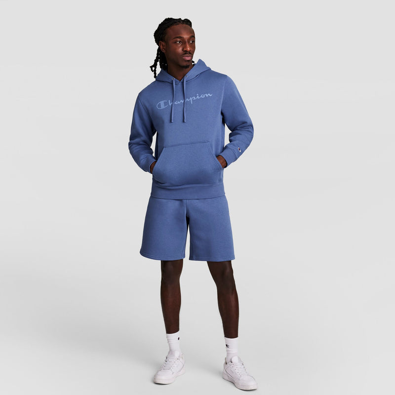 A man stands against a plain background wearing a blue Champion Powerblend Hoodie with a tonal script logo, matching fleece shorts, and white sneakers, hands in his hoodie pocket, looking slightly to the side.