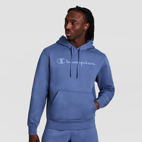 A man wears a blue Champion Powerblend Hoodie with a tonal script logo and matching fleece pants, standing against a plain light background, looking left with one hand in his pocket.